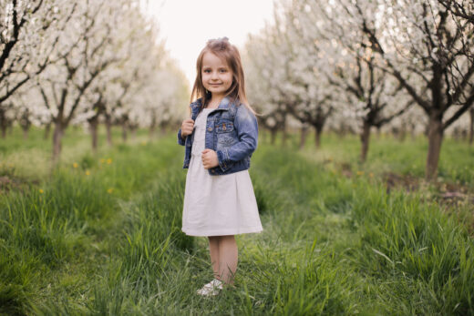 criança menina usando vestido branco com jaqueta jeans sobreposta em um campo arborizado e gramado